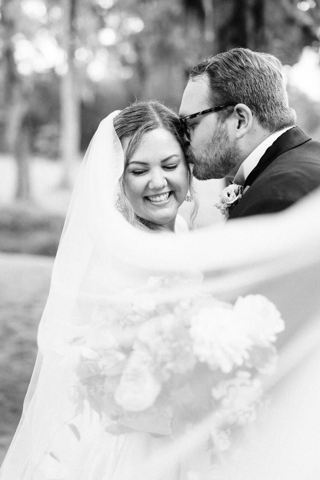 A groom lovingly kisses his bride's forehead in a candid, black and white wedding photo