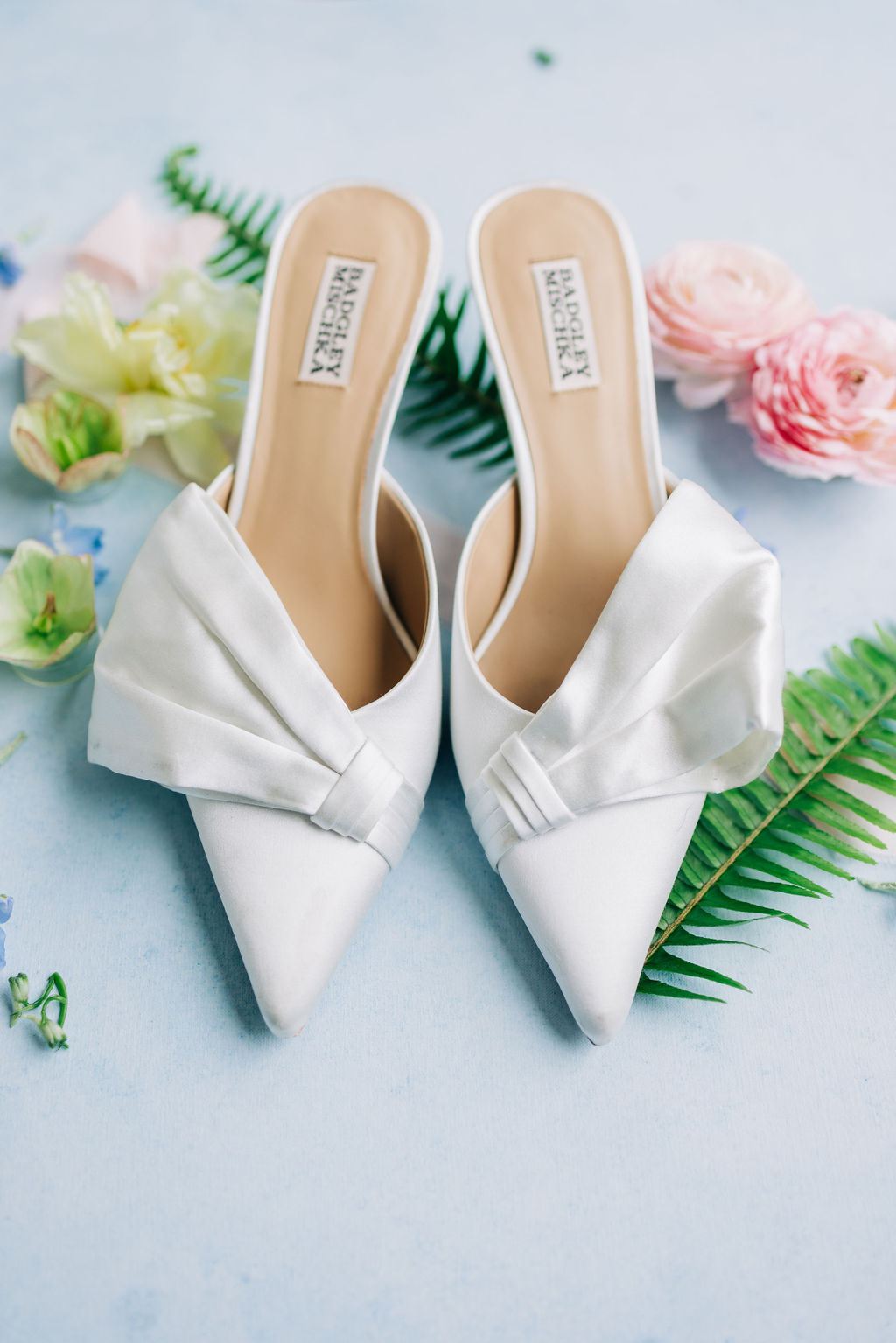 A pair of white Badgley Mischka bridal mules with bow detailing rests on a light blue background, surrounded by flowers and green ferns
