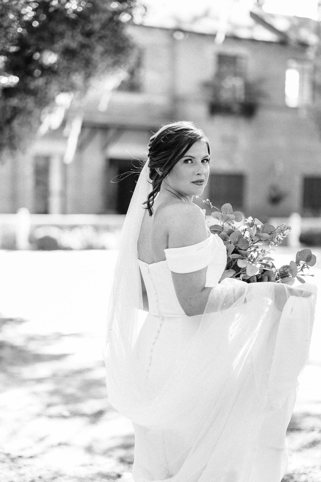 A bride in a white dress holds a bouquet of flowers and smiles over her shoulder at the camera in a sun-dappled outdoor setting.