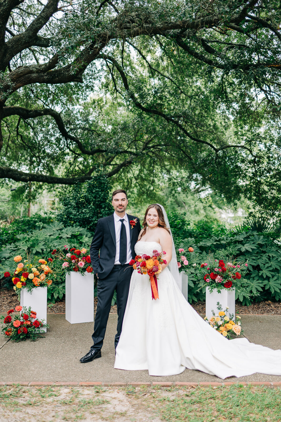Bride and groom stand surrounded by vibrant red, orange, and yellow floral arrangements