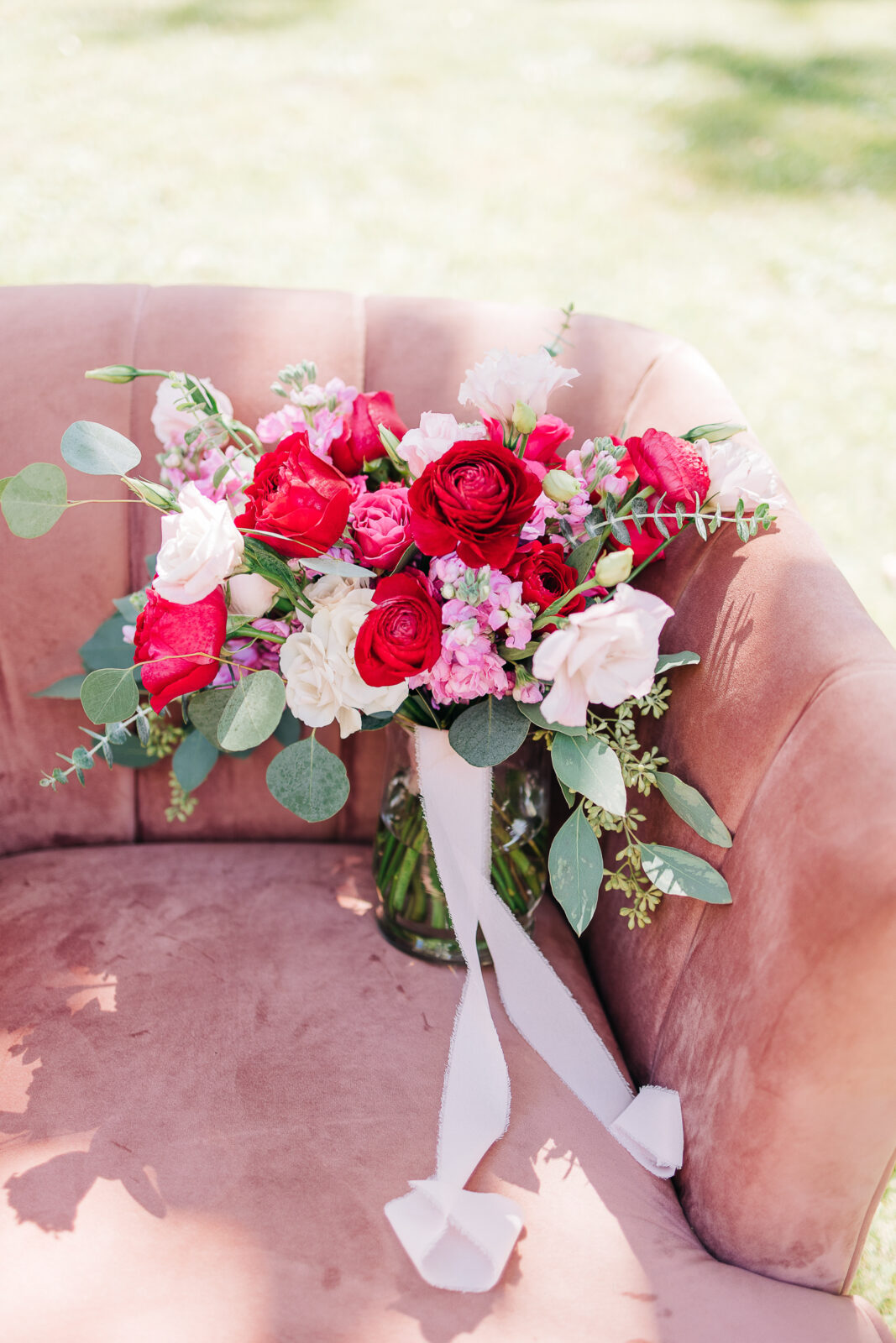 Bright bridal bouquet of red roses, pink flowers, and eucalyptus greenery on a blush velvet chair for a wedding in Columbia, SC