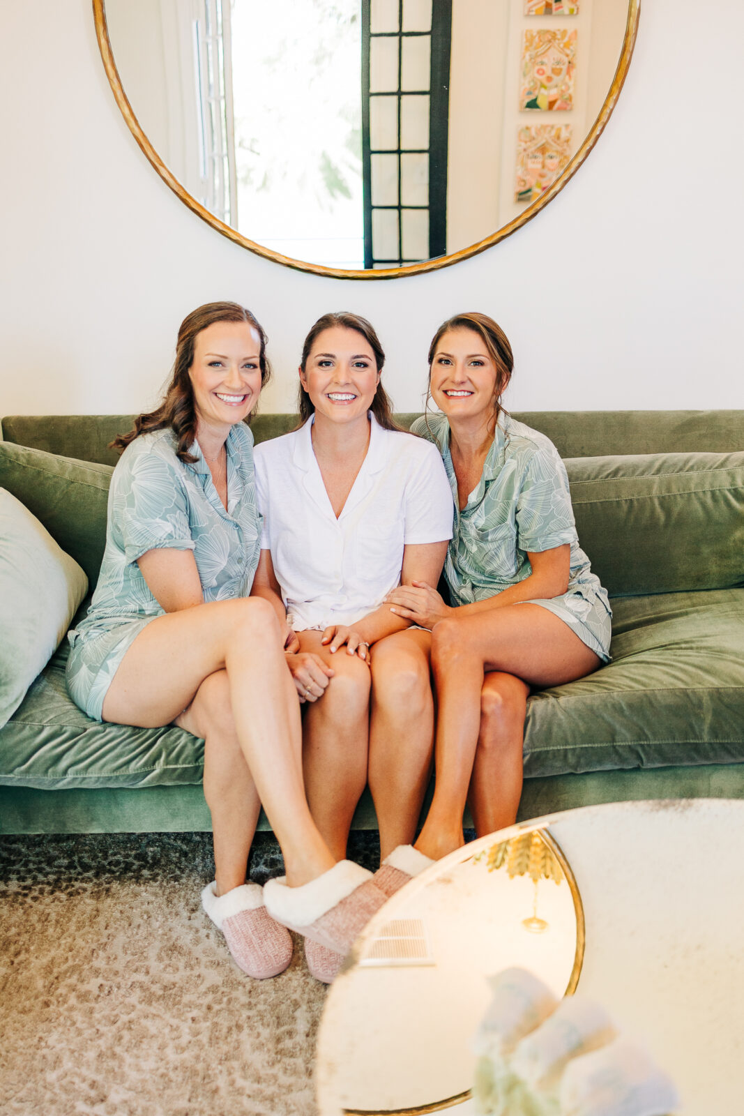 Bride and two bridesmaids wearing matching pajamas and slippers, sitting on a green velvet couch during wedding day preparations