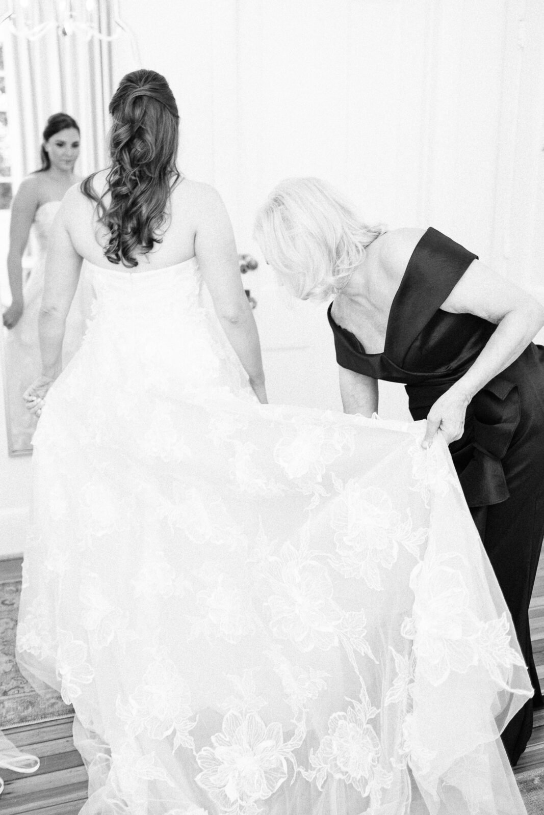 Black and white photo of the mother of the bride helping adjust the train of the lace wedding dress