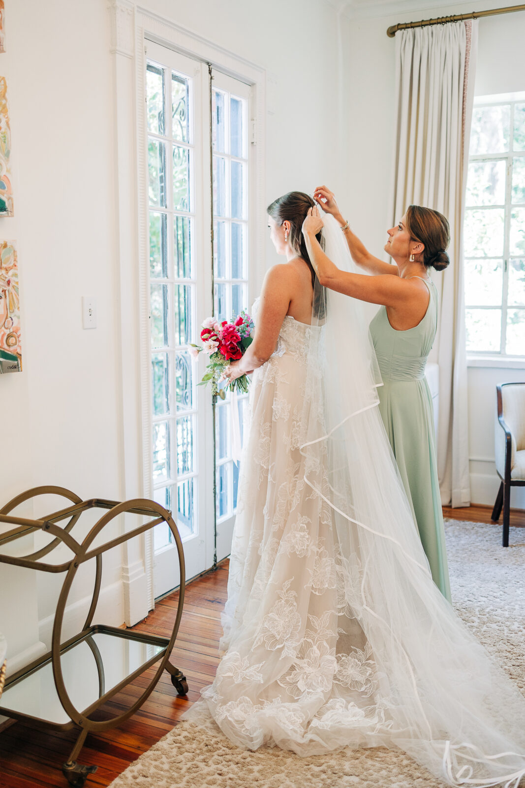 A bridesmaid in a pale green dress carefully placing the wedding veil on the bride near french doors with natural light