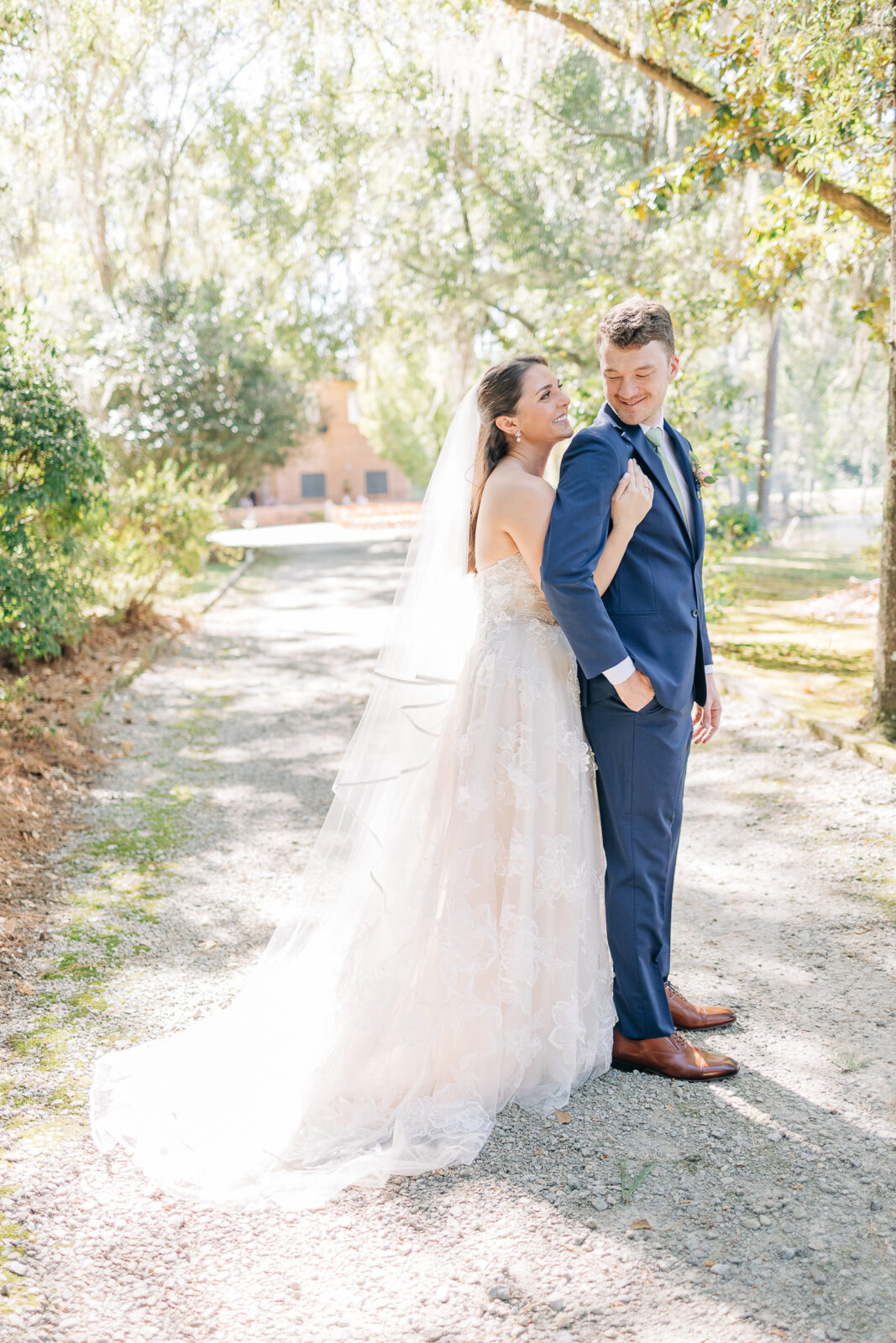 Full-length portrait of the bride in a long veil and lace gown with the groom in a navy suit and green tie outdoors