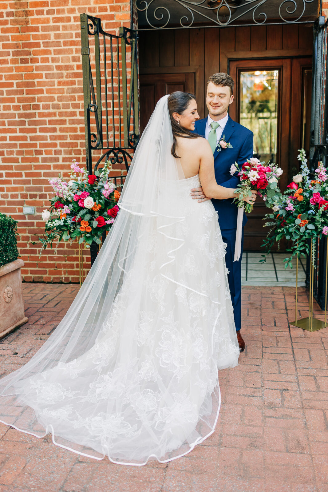 Smiling bride and groom embracing in their first look, showing the back of the bride's long lace wedding dress and veil