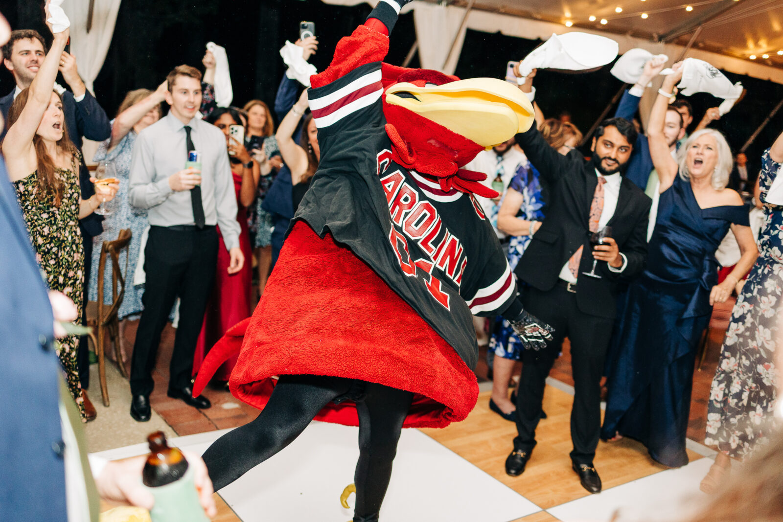 Enthusiastic wedding guests raising napkins and dancing with the Gamecock mascot at a South Carolina wedding reception