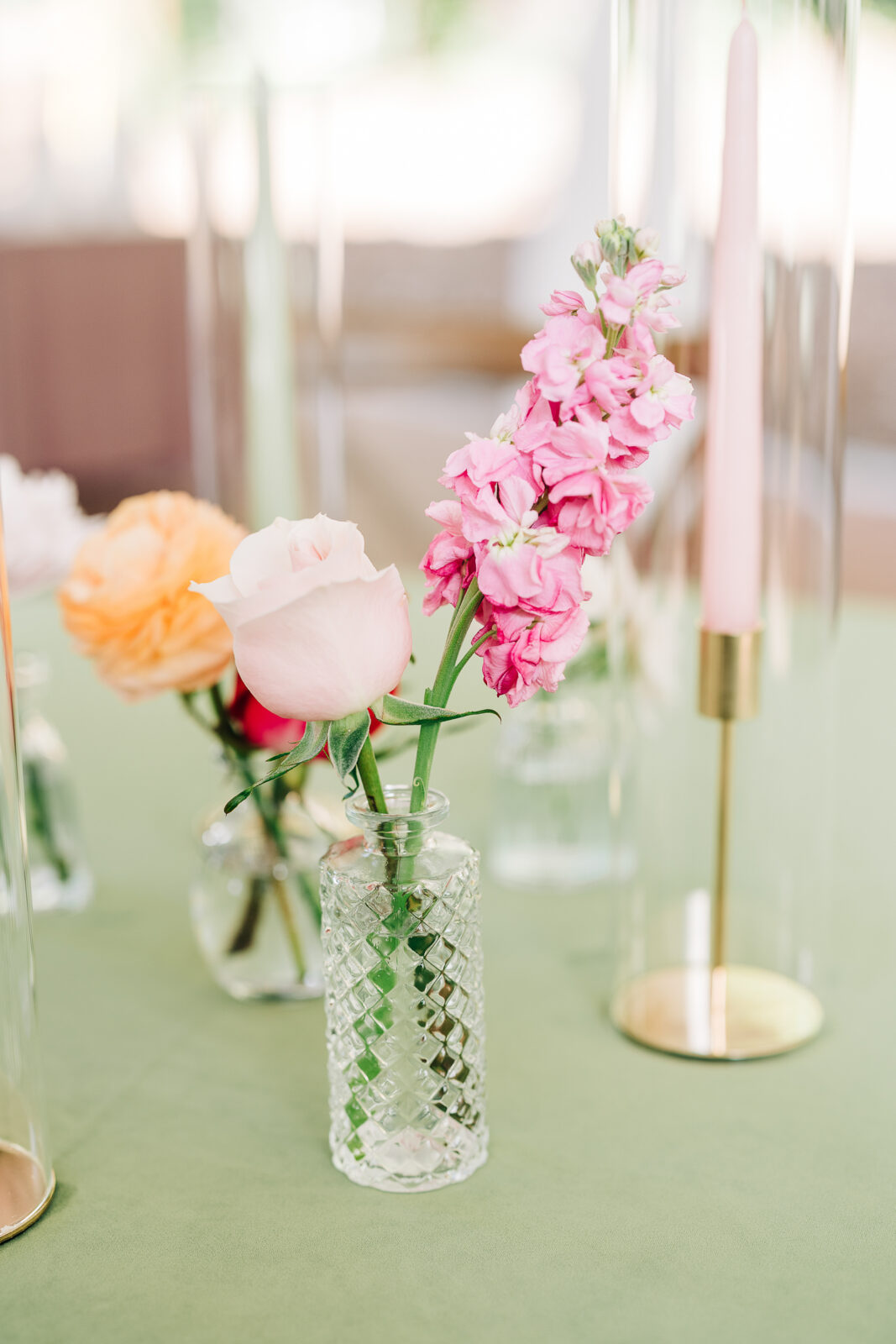 Close-up wedding centerpiece detail with pink stock, orange ranunculus, and pink taper candles on a green table