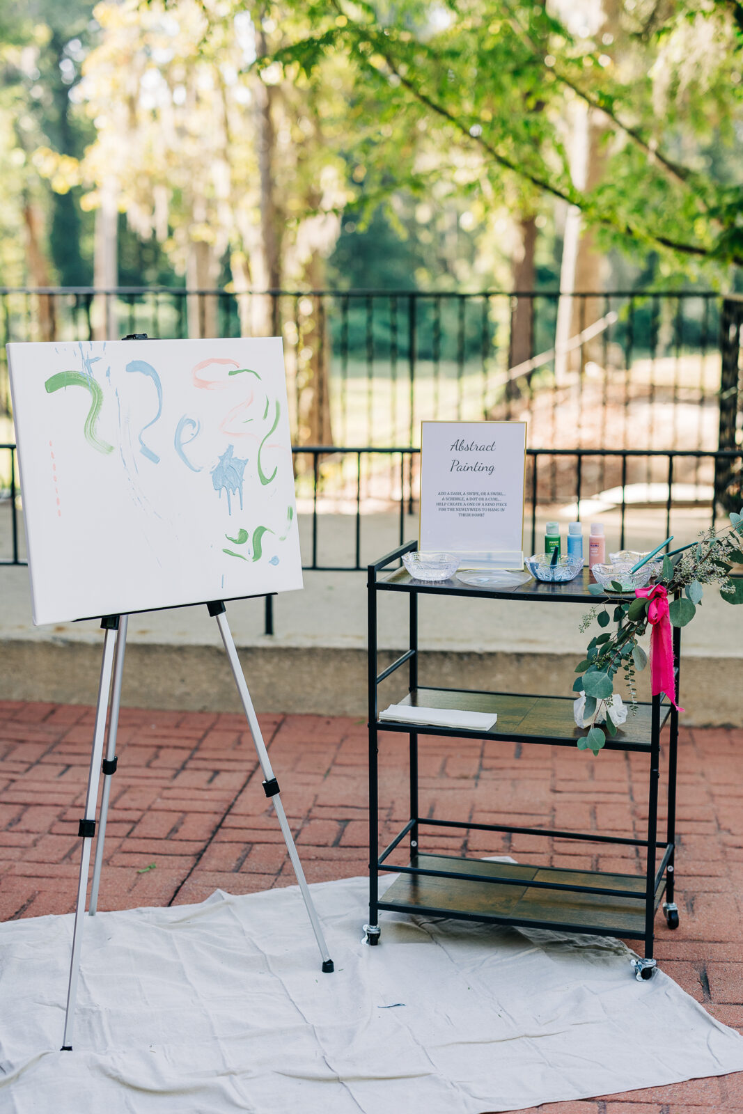 An outdoor wedding activity station with an easel holding an abstract painted canvas and a cart displaying paints and a sign for "Abstract Painting