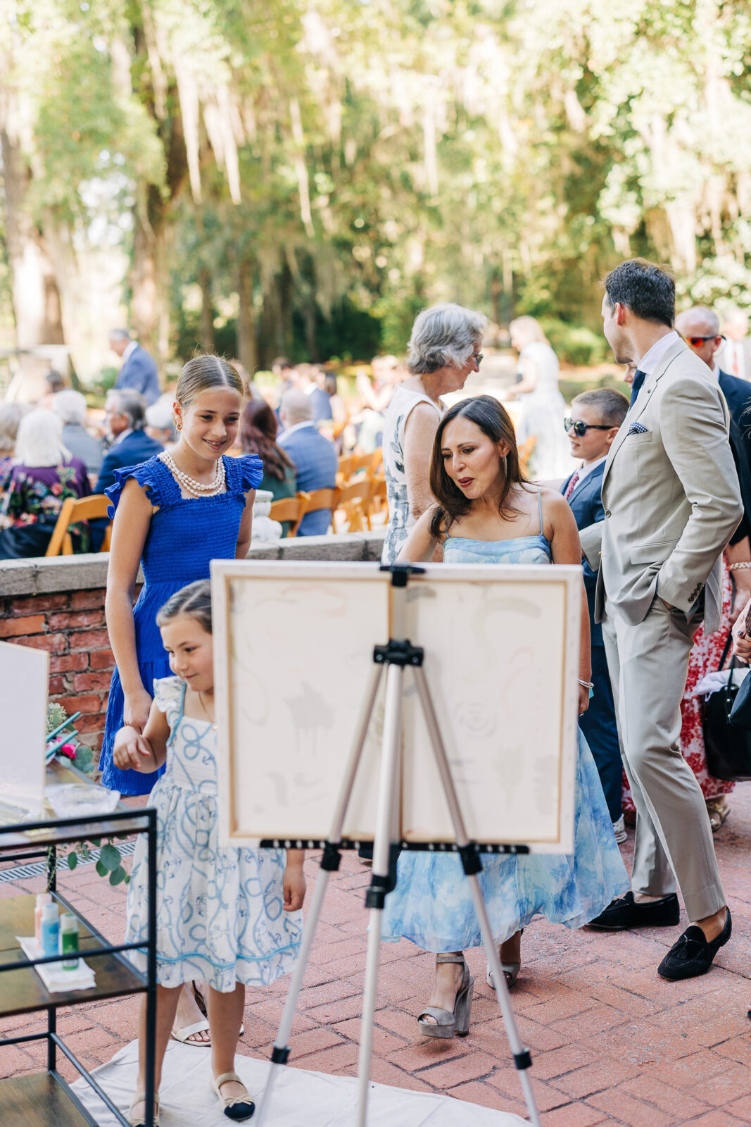 Wedding guests, including a woman in a blue dress and two girls, look at a canvas on an easel set up on a brick patio during an outdoor wedding reception