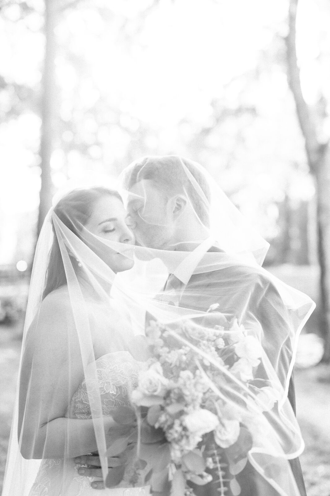 Bride and groom veil kiss under the trees in black and white at The Millstone at Adams Pond wedding venue