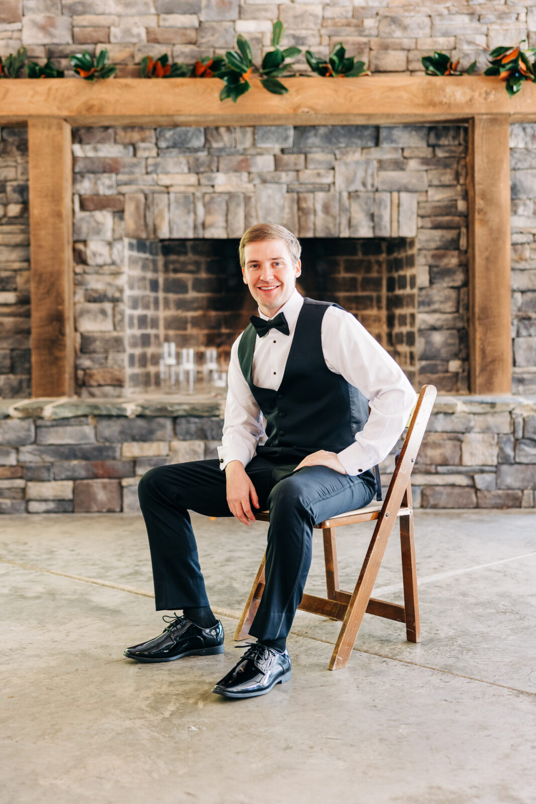Groom smiling in black tuxedo vest sitting in front of stone fireplace at Oakbrook Farms Wedding South Carolina