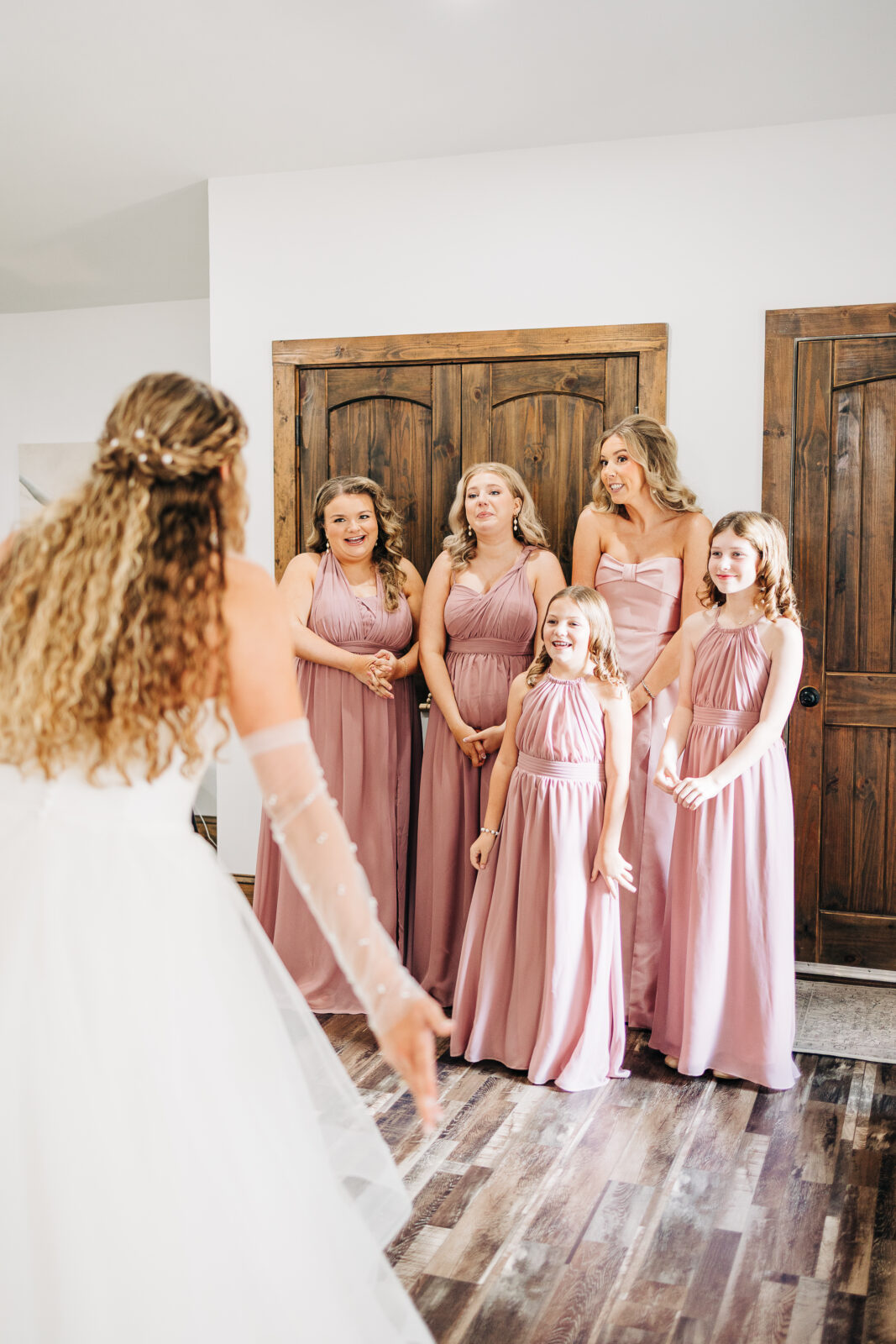 Bride showing off her dress to bridesmaids in rose-colored gowns in a first look moment
