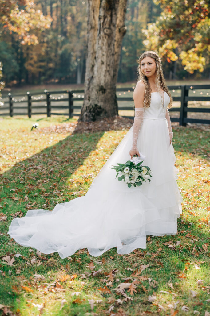 Bride standing in fall foliage with white dress and wooden fence at Oakbrook Farms Southern Wedding