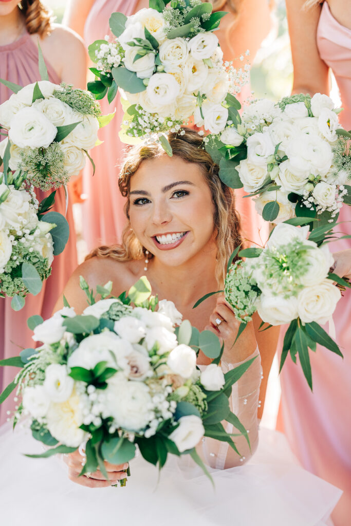 Smiling bride surrounded by beautiful white and green bouquets and bridesmaids in pink dresses
