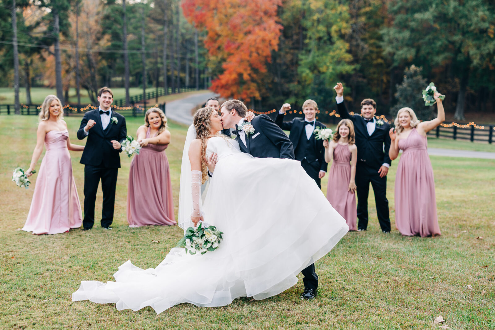 Bride and groom dipping and kissing with cheering wedding party in grassy field