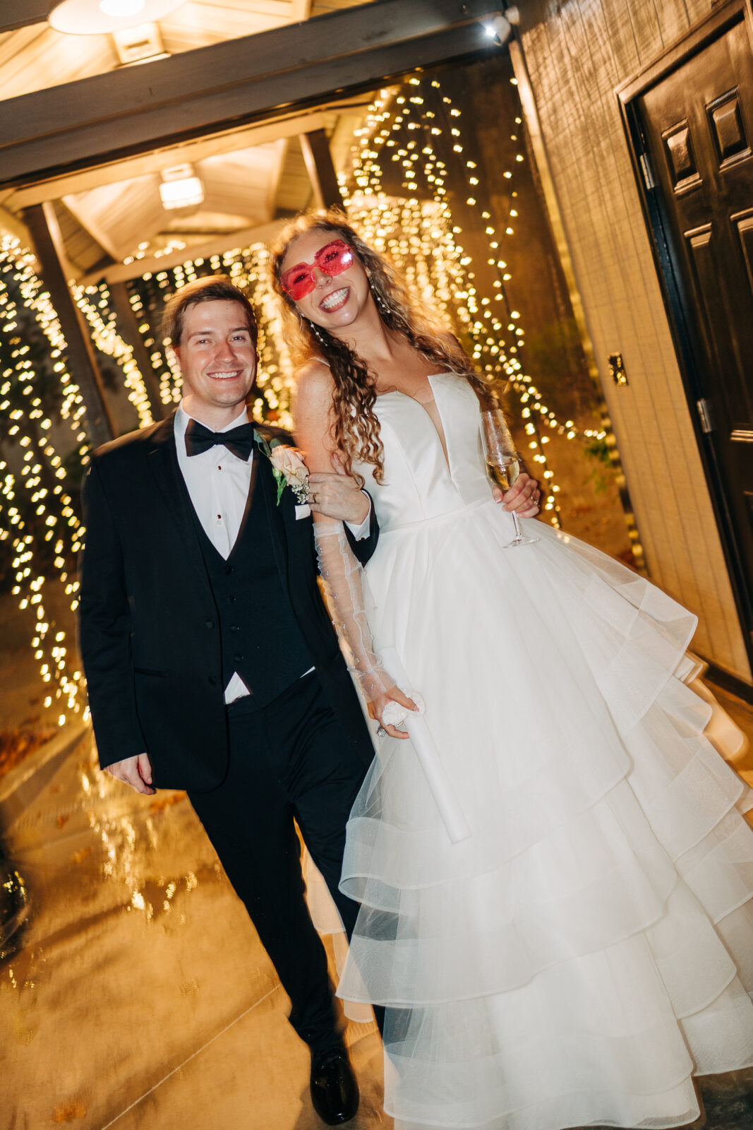 Bride wearing fun pink heart glasses posing with groom under romantic string lights