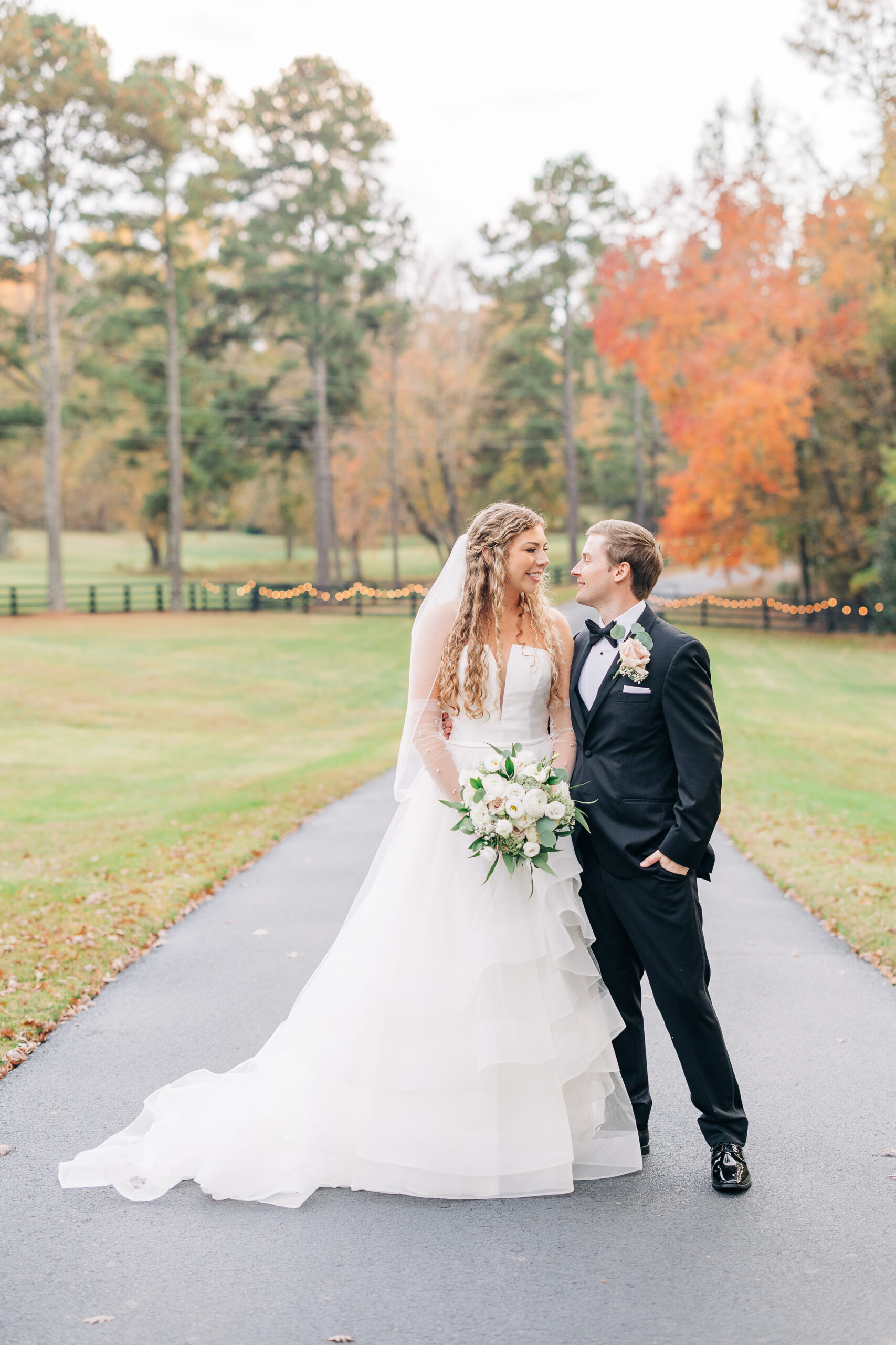 Bride and groom posing and looking at each other on a paved path with fall foliage in the background