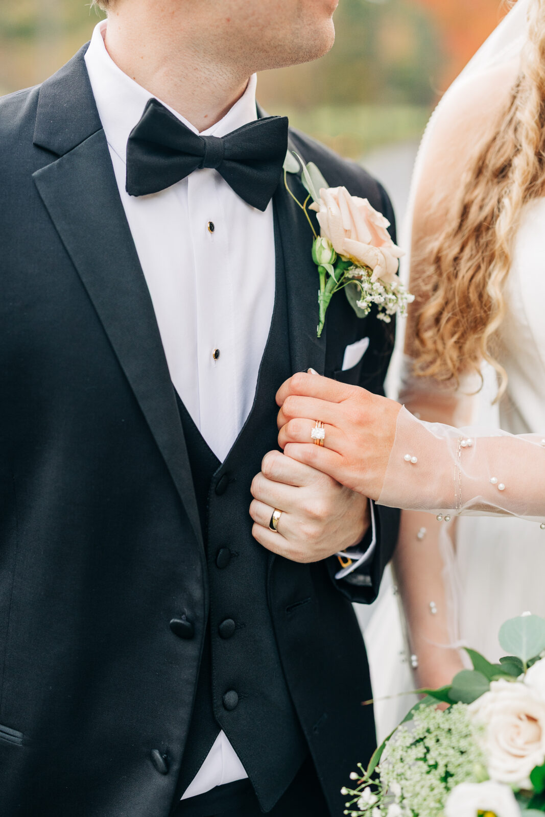Close-up of the couple's hands; bride's hand with pearl-detailed sleeve holding the groom's tuxedo lapel, showing wedding bands