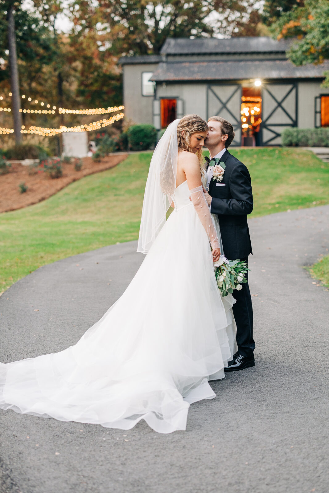 Groom kissing bride on cheek with long train outside the barn venue at Oakbrook Farms Wedding South Carolina