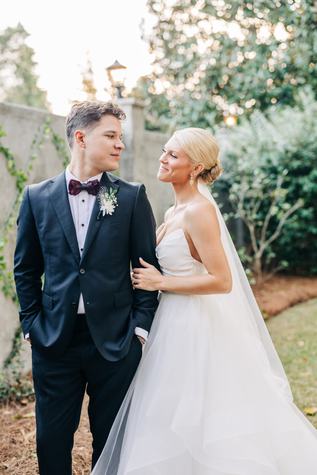 Full-length portrait of the bride and groom standing close together, with the bride's train flowing behind her, against a concrete, vine-covered wall at the Hartley House Wedding South Carolina