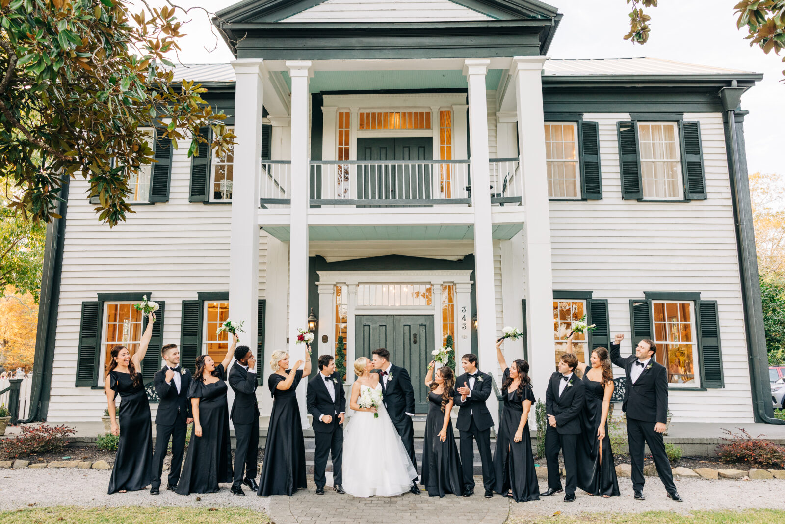 Wide shot of the entire wedding party in black gowns and tuxedos raising their bouquets and hands in a cheer outside the grand white southern colonial-style house at the Hartley House Wedding South Carolina