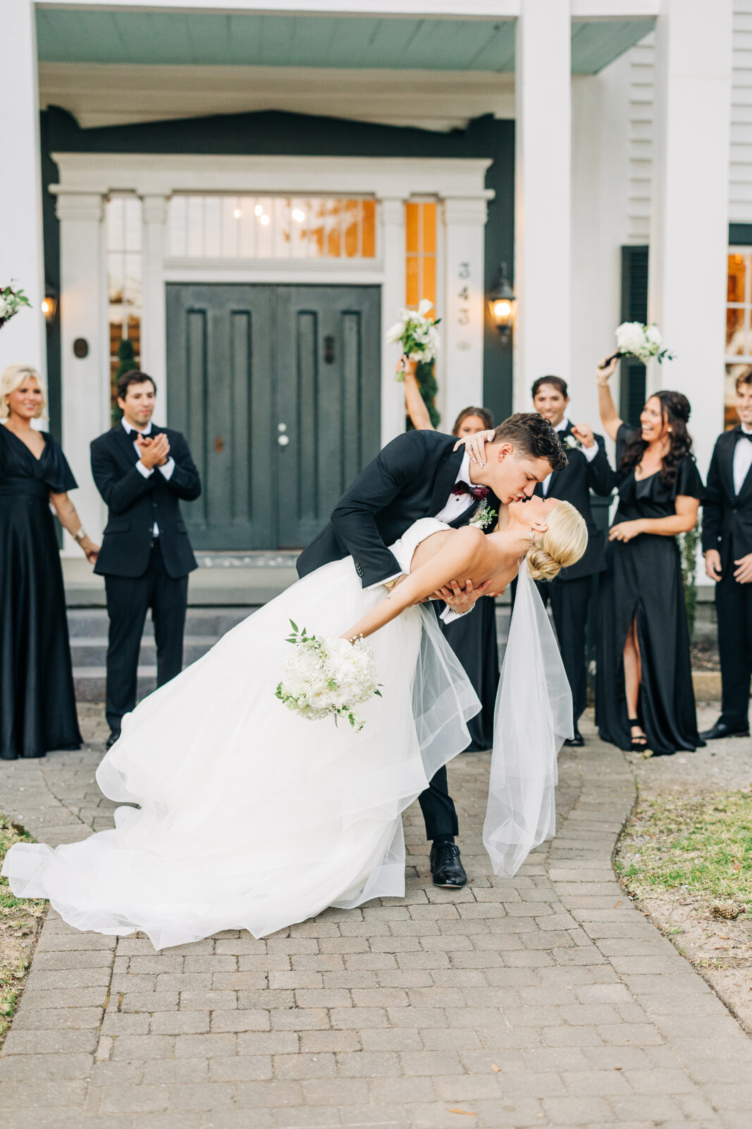The groom dips the bride for a kiss on the brick walkway, surrounded by their smiling wedding party in black attire, in front of the venue's entrance