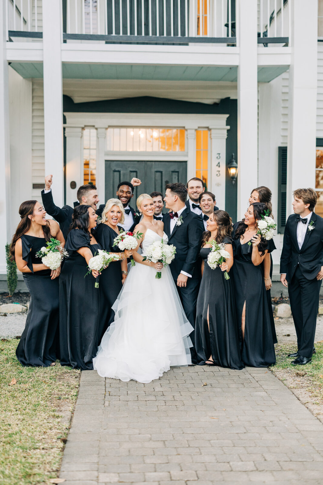 Group portrait of the bride and groom smiling with their wedding party in black dresses and tuxedos in front of the house entrance, captured at the Hartley House