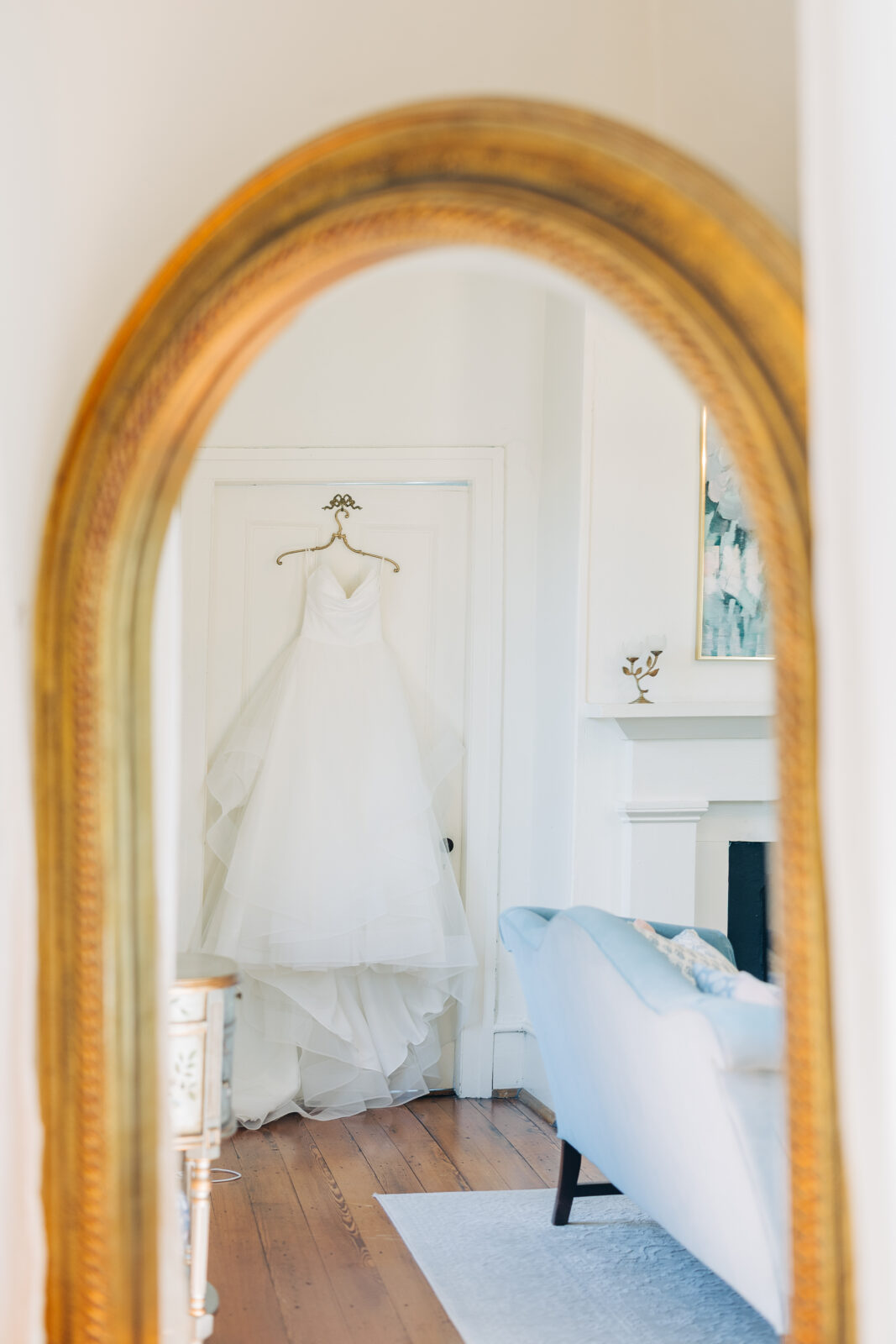 A reflection of the white strapless wedding dress hanging on a door in a bright, elegant room with a blue couch, captured in a large ornate gold mirror at the Hartley House Wedding South Carolina