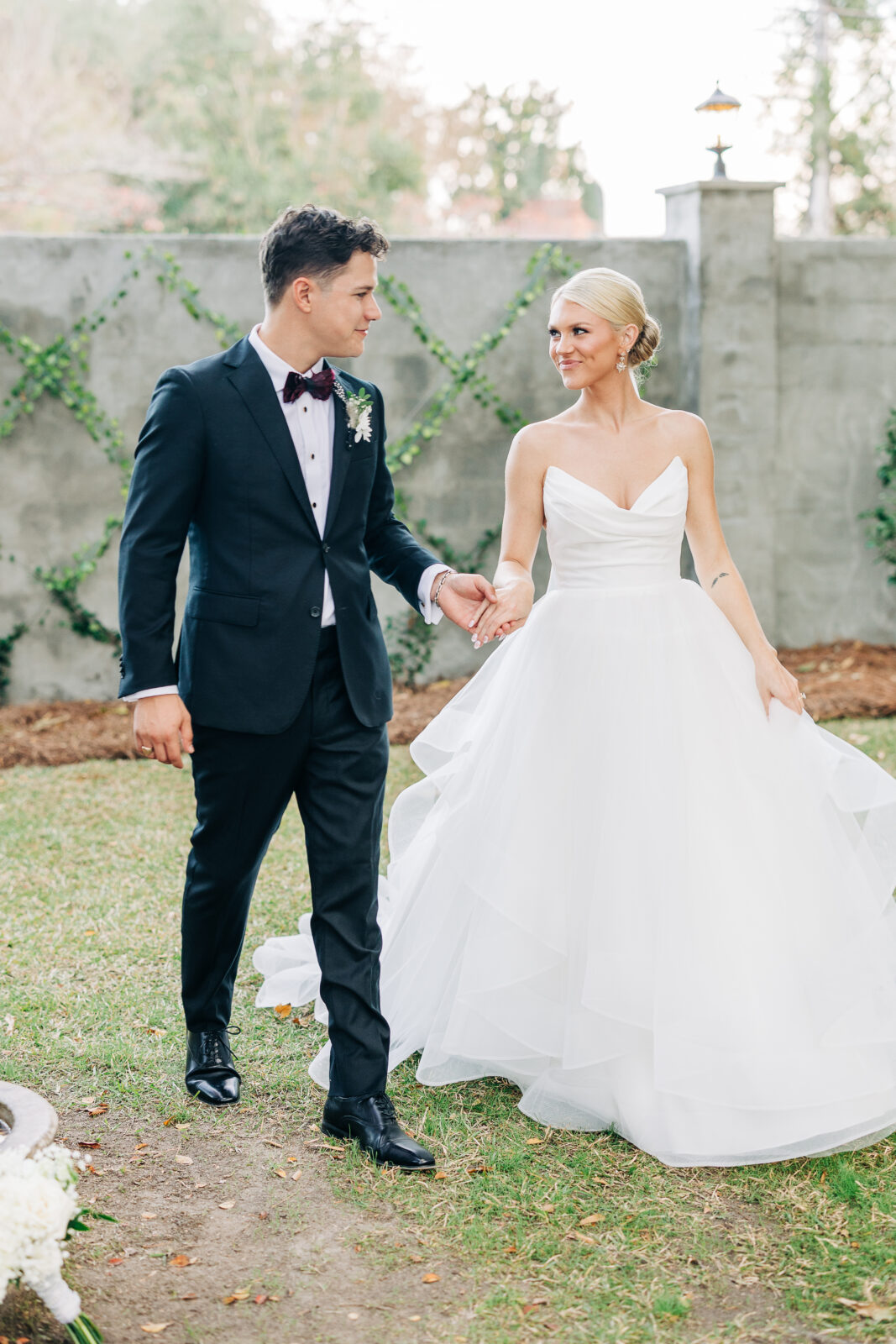The bride and groom smiling at each other while walking hand-in-hand across a grassy area near a cement wall after their Hartley House Wedding South Carolina ceremony.