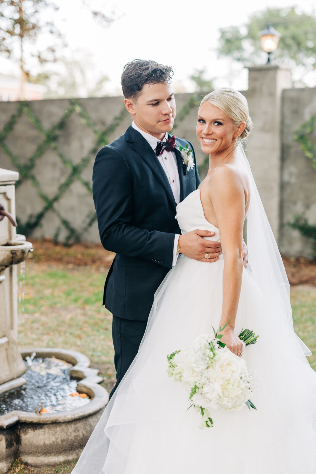 A portrait of the smiling newlywed couple standing beside a stone fountain in the courtyard of the Hartley House Wedding South Carolina venue