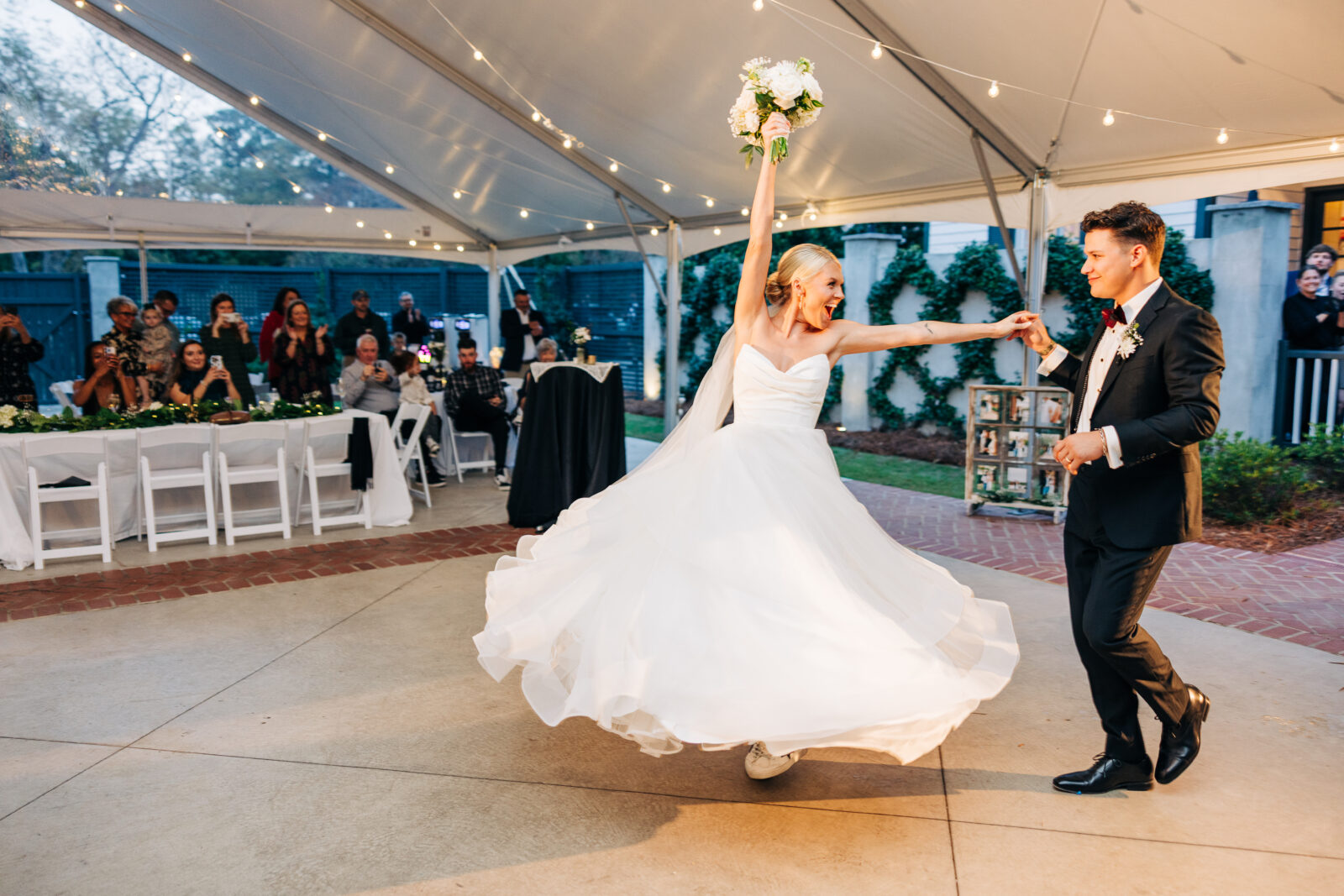 The bride, holding her bouquet high and twirling in her tiered gown, and the groom dance happily during the evening reception under the tent at the Hartley House Wedding South Carolina.