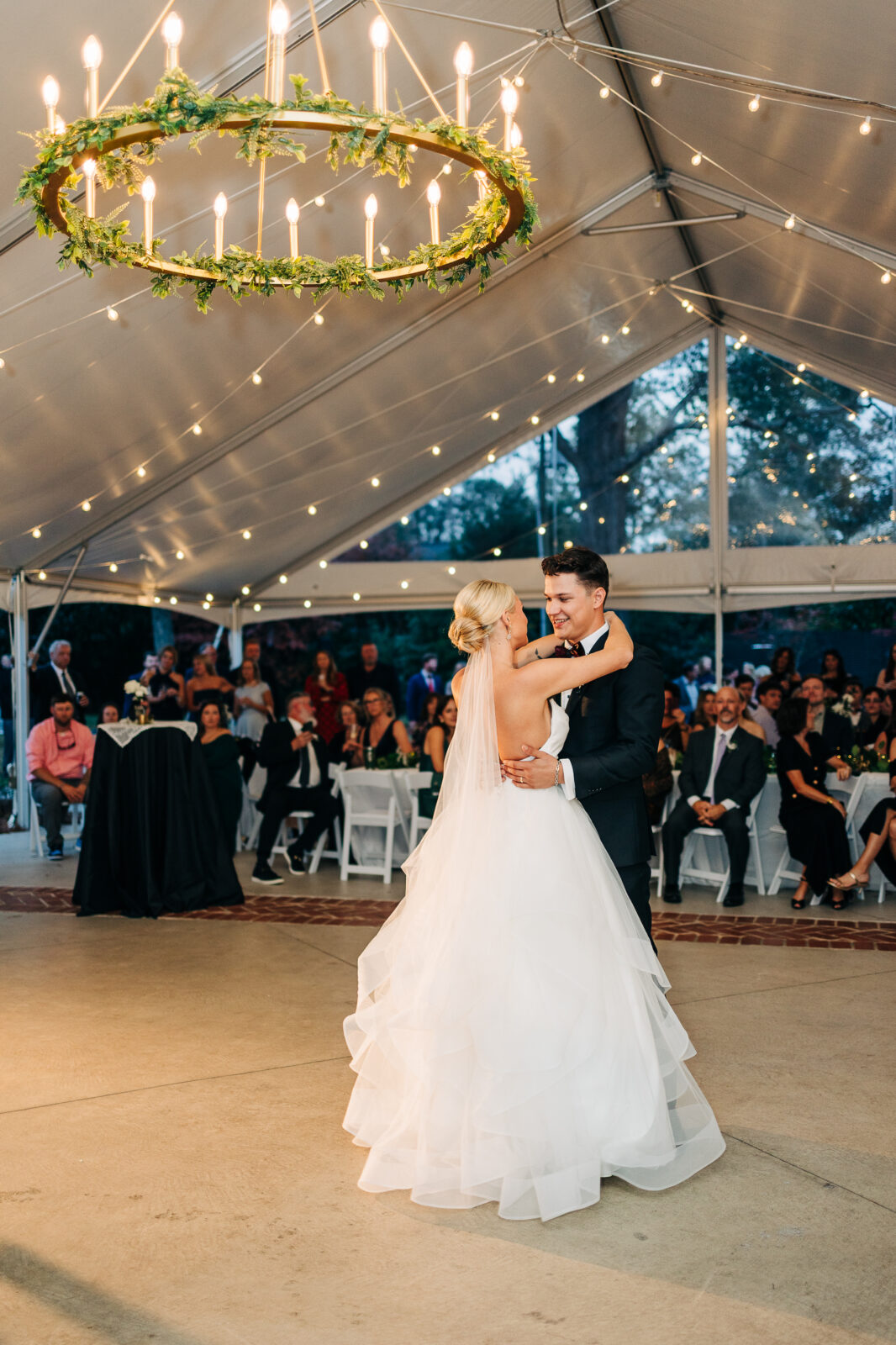 The bride and groom share a loving moment during their first dance under the beautifully lit tent and garland chandelier at the Hartley House Wedding South Carolina reception