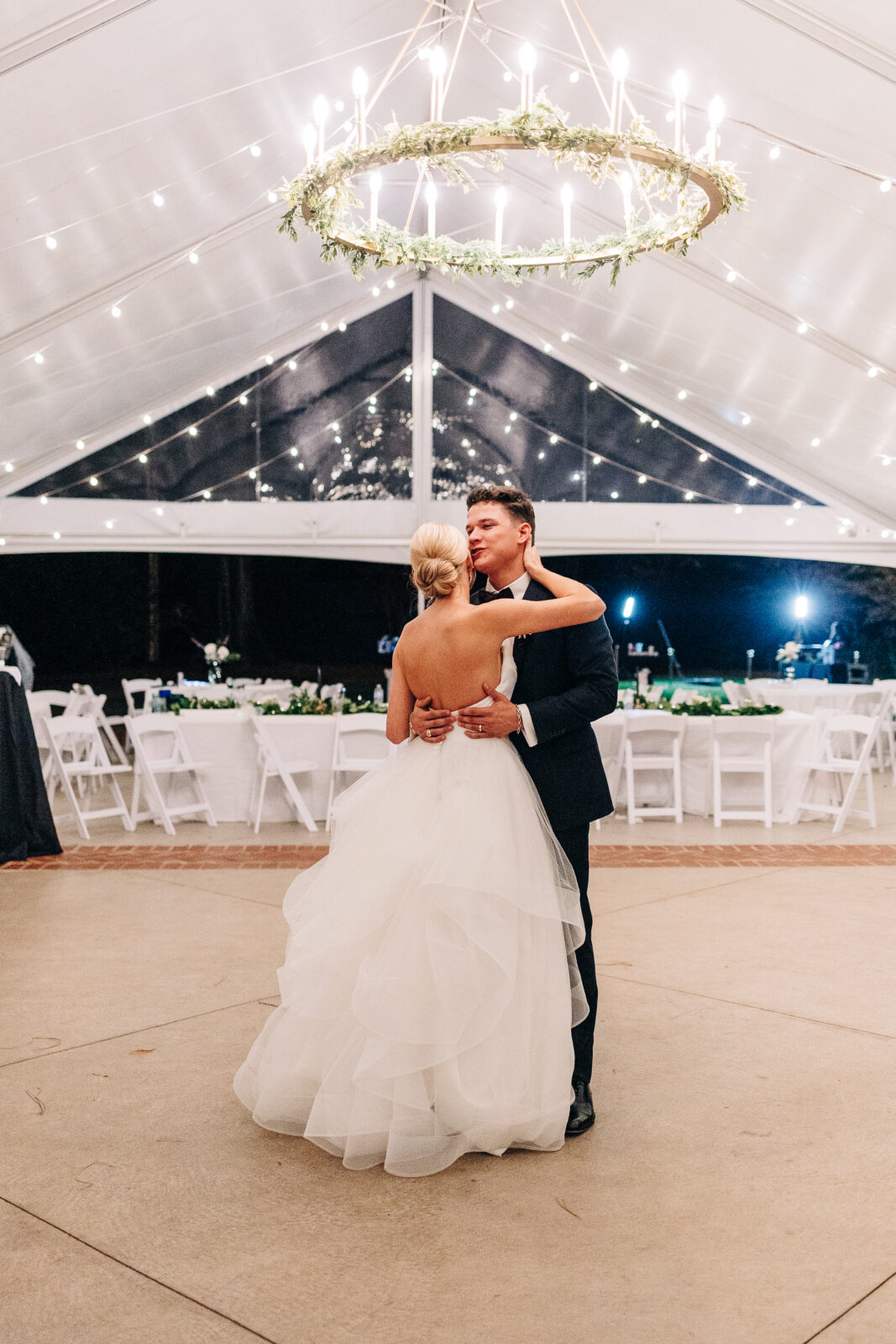 Over-the-shoulder view of the bride and groom kissing during their first dance, showing the tiered back of the bride's dress and the elegant tent ceiling