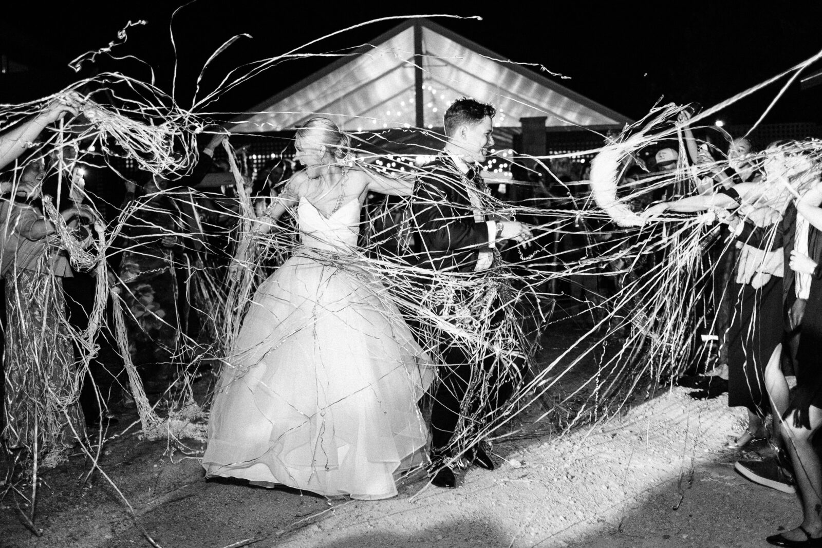 The jubilant bride and groom running through a tunnel of guests and strands of golden silly string during their grand exit at the night-time Hartley House Wedding South Carolina reception