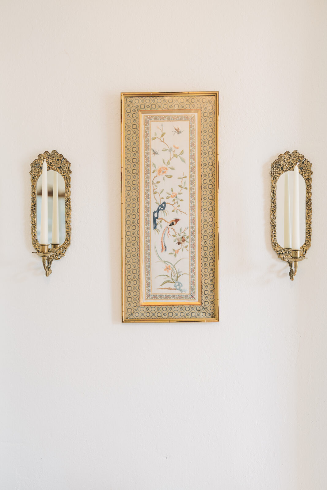 Interior decor detail showing a long, framed piece of art featuring birds and flowers, flanked by two ornate gold sconces with pillar candles on a white wall at the Hartley House Wedding South Carolina