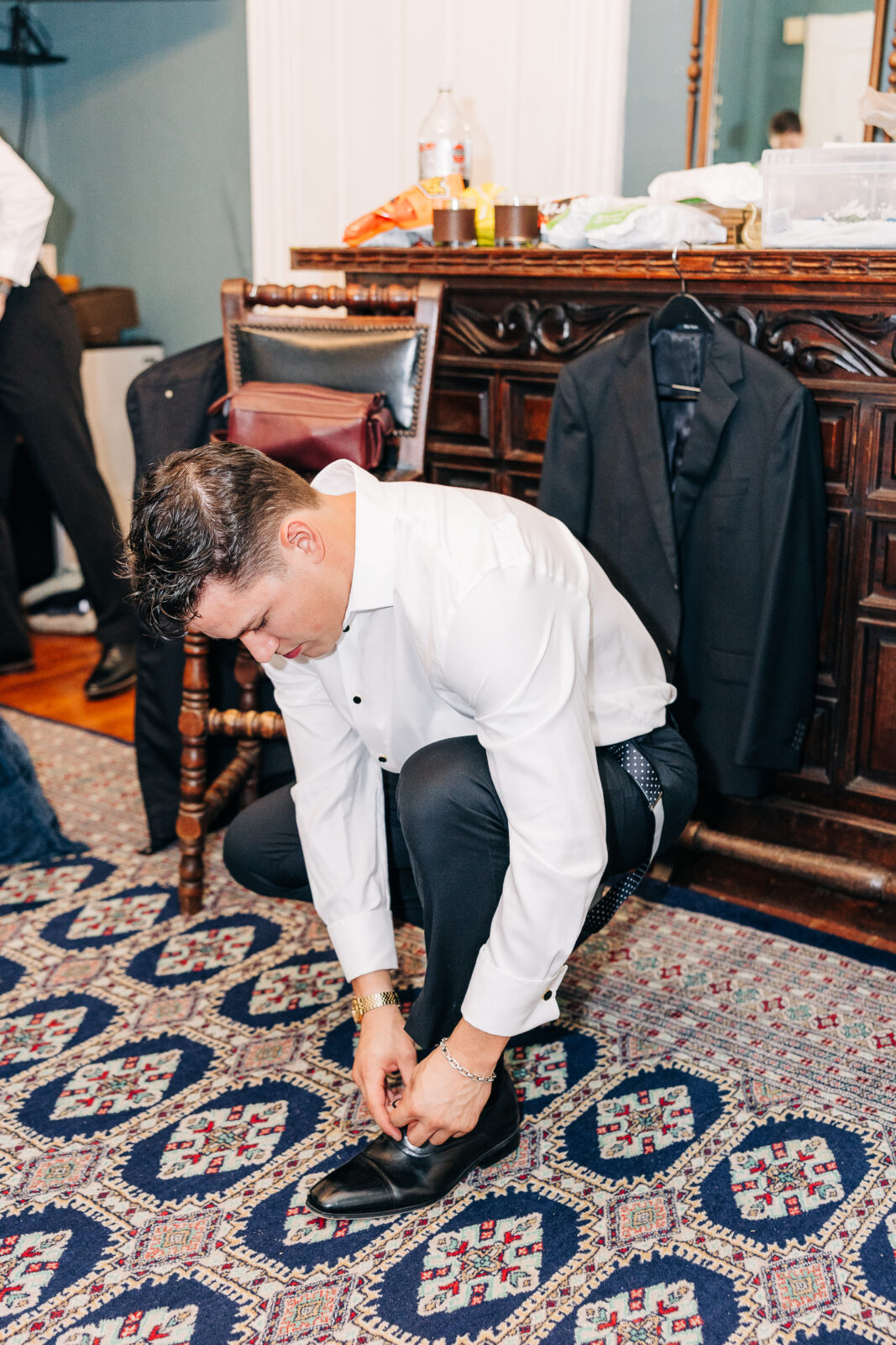 The groom in a white shirt and black trousers bending down to tie his black leather shoes on a patterned rug during his preparation for the Hartley House Wedding South Carolina