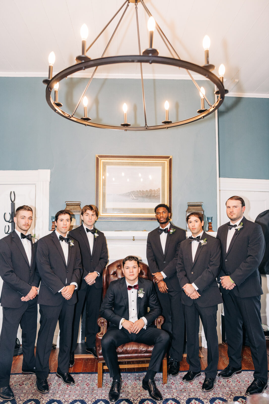 The groom in a black tuxedo sits reading a heartfelt card from the bride on a tufted chair in the preparation room before the Hartley House Wedding South Carolina ceremony