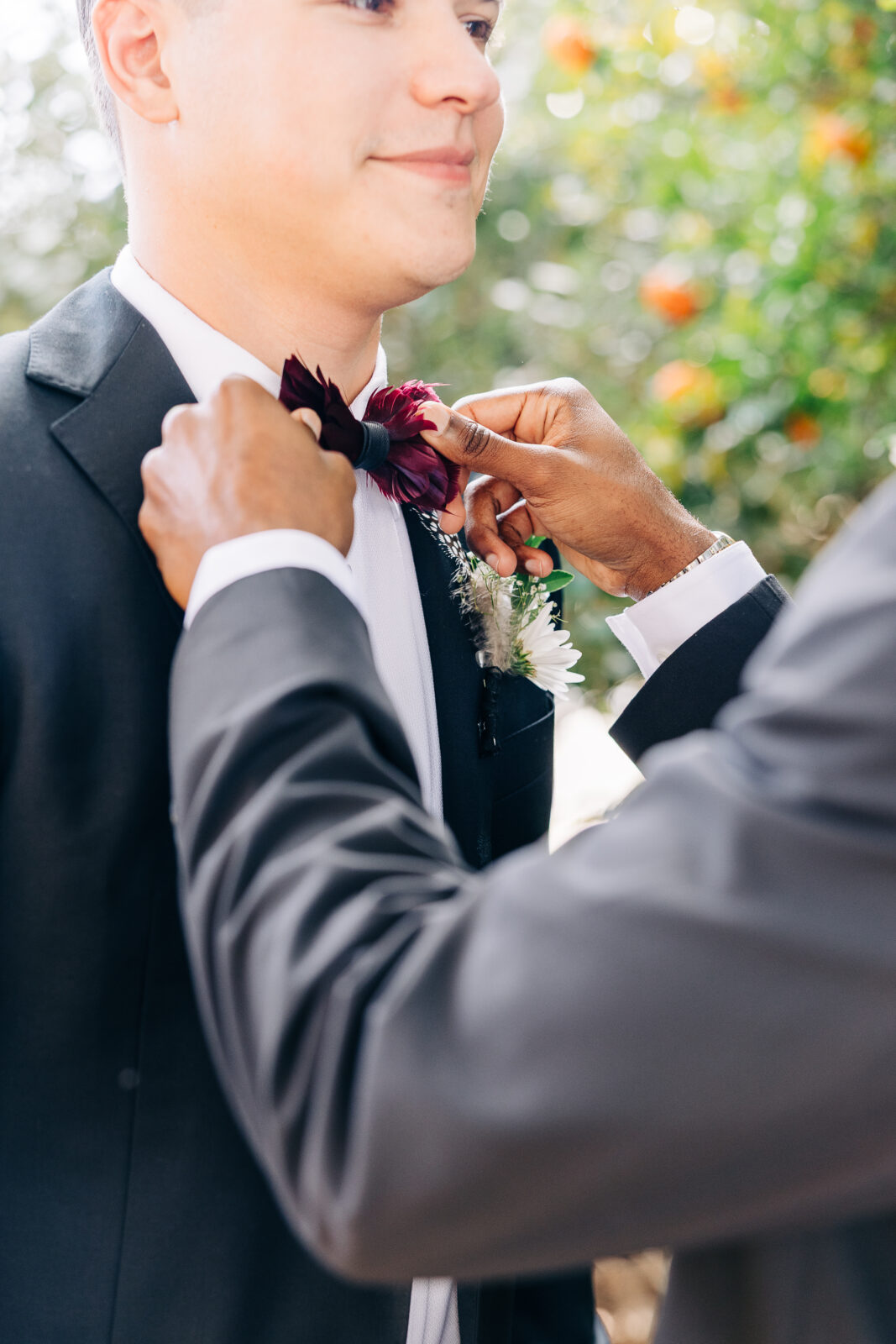 Close-up outdoors shot of a groomsman adjusting the groom's deep red velvet bowtie and white flower boutonnière for the Hartley House Wedding South Carolina