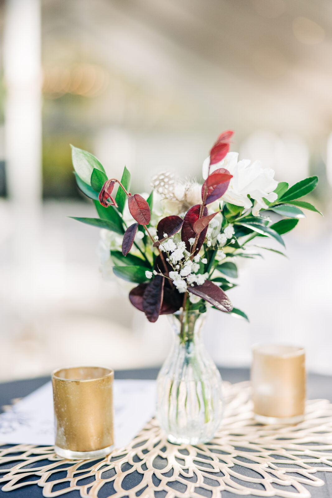 Centerpiece detail showing a small glass vase holding a floral arrangement with white blooms, dark red leaves, and feathers, sitting on a gold filigree mat at the Hartley House Wedding South Carolina reception