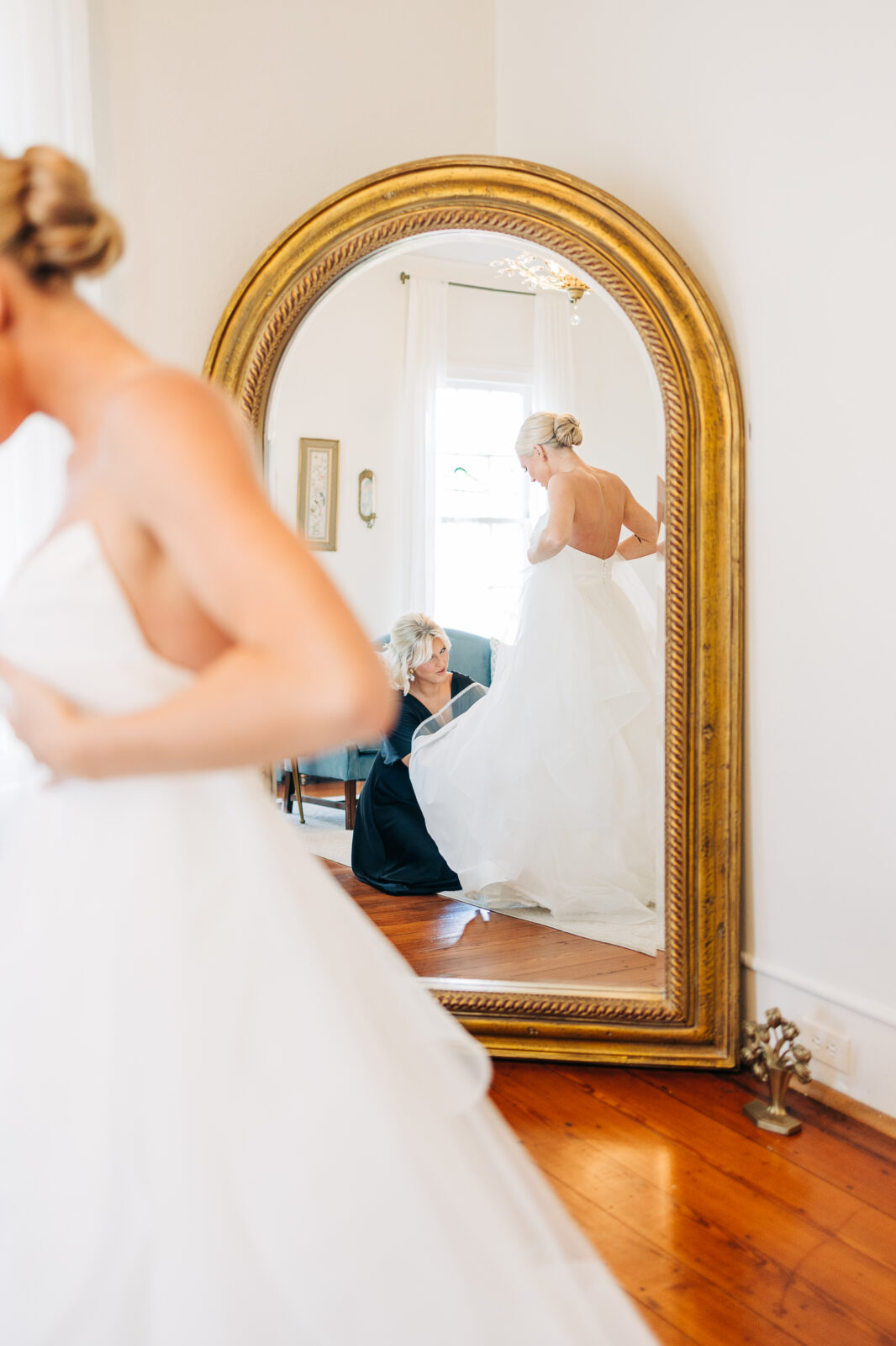 A reflection in a large gold mirror showing the bride in her white wedding dress being assisted by an older woman in a black dress in the bridal suite at the Hartley House Wedding South Carolina