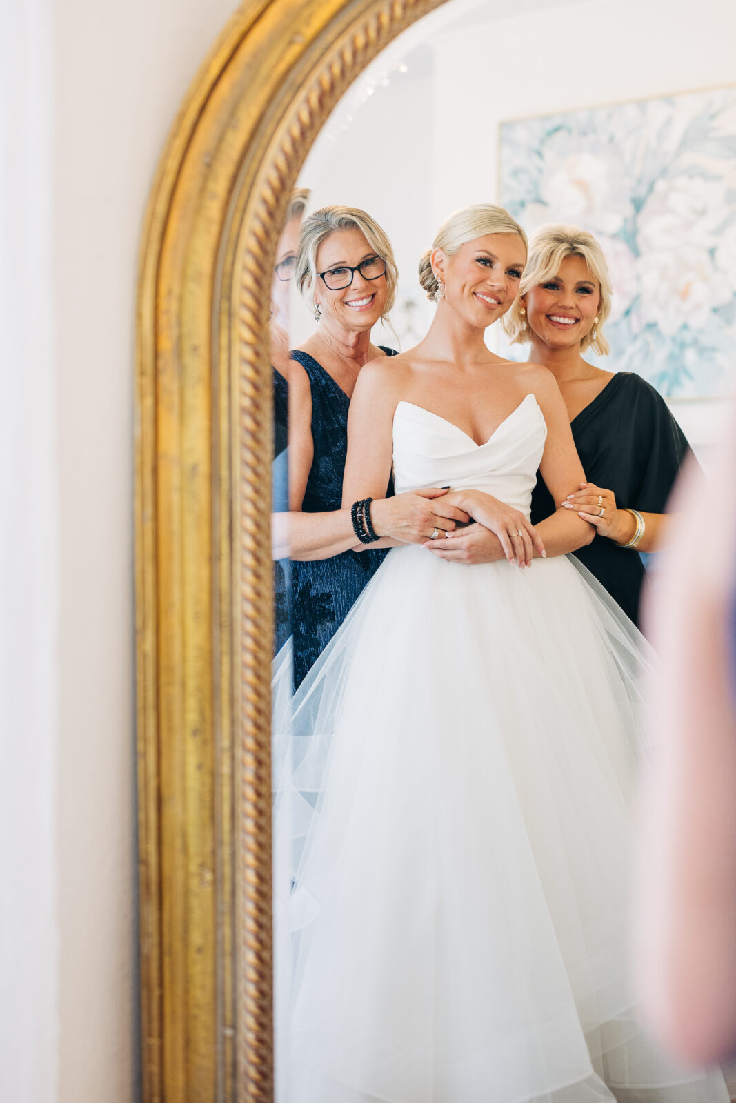 The bride, wearing a strapless gown, poses in a gold-framed mirror with two older women (the mothers), all smiling happily on her wedding day at the Hartley House Wedding South Carolina