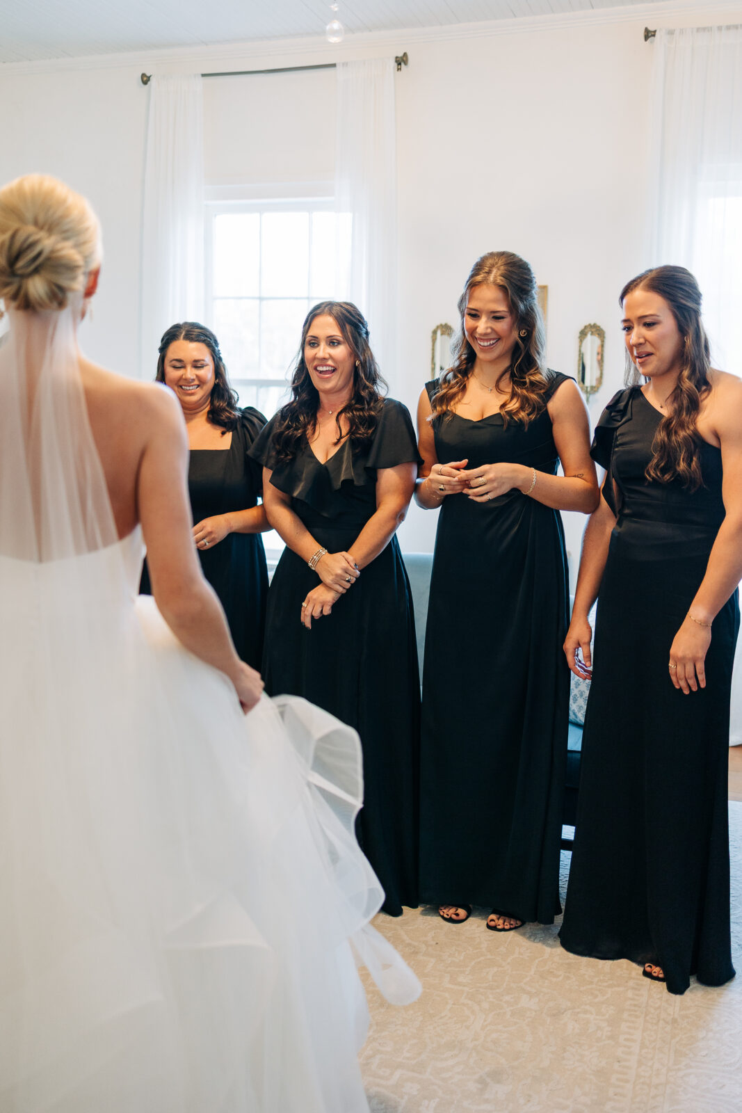 The bride, dressed in her white gown and veil, stands with her back to the camera, showing off her dress to her four bridesmaids who are wearing elegant black dresses at the Hartley House Wedding South Carolina