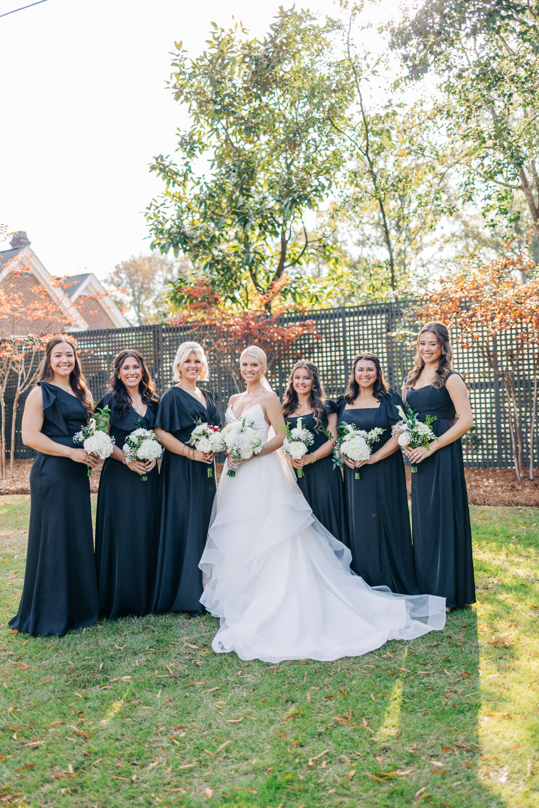 The bride in her strapless white tiered gown stands with her five bridesmaids in long black dresses, posing for a group portrait on a lawn surrounded by trees at the Hartley House Wedding South Carolina