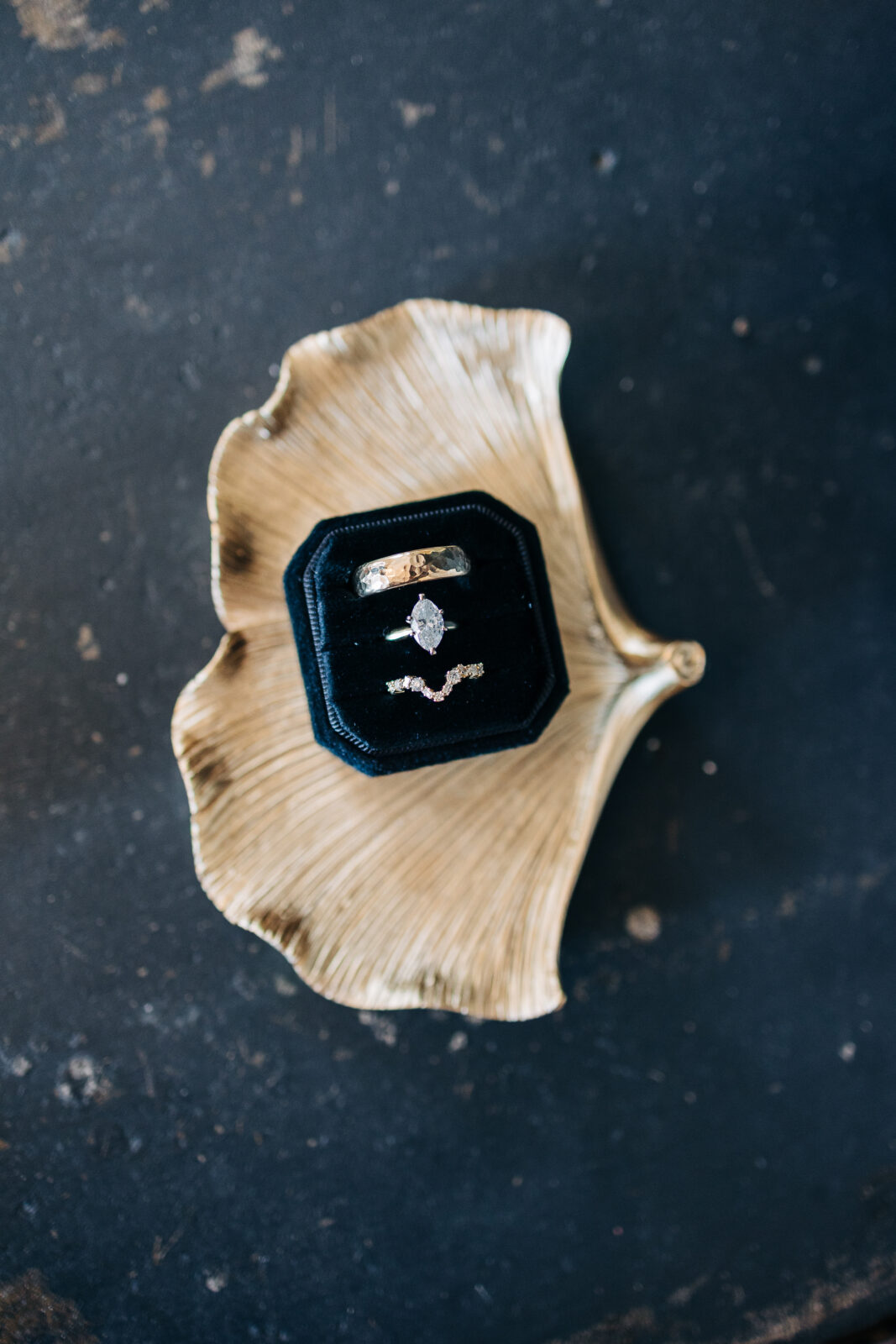 Close-up of the bride's diamond engagement ring, matching curved wedding band, and the groom's gold wedding band resting in a velvet box on a gold ginkgo leaf dish at the Hartley House Wedding South Carolina