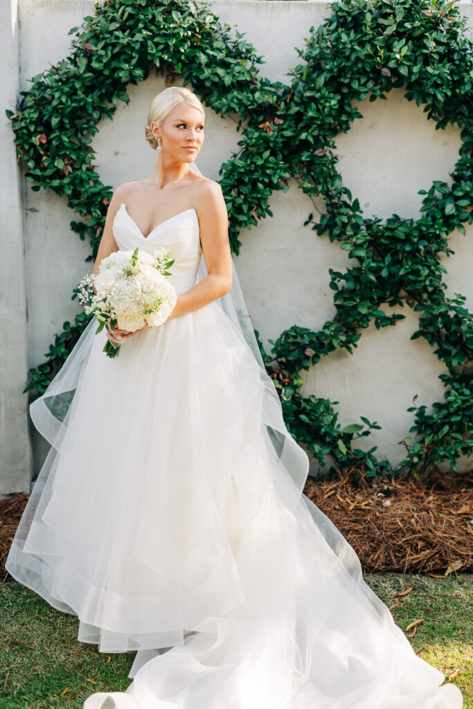 Full-length portrait of the blonde bride in her strapless tiered gown and veil, posing against a white wall decorated with large green ivy wreaths at the Hartley House Wedding South Carolina