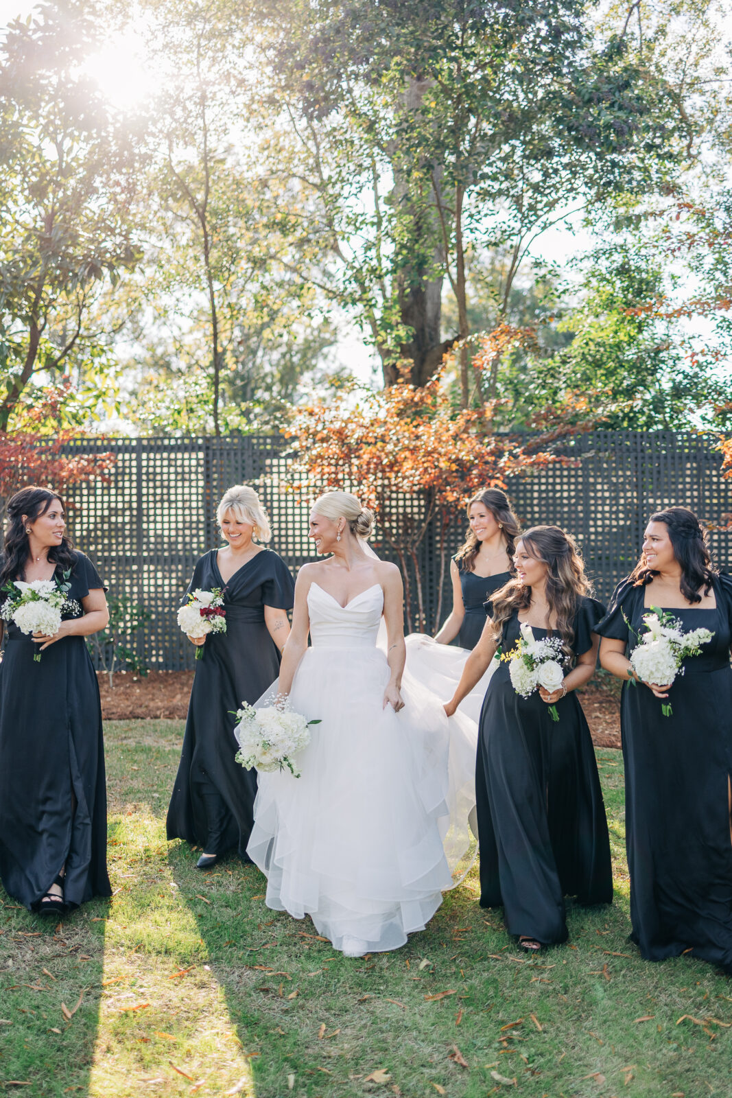 The bride and her five bridesmaids in long black dresses walking across a sunny grassy lawn outside