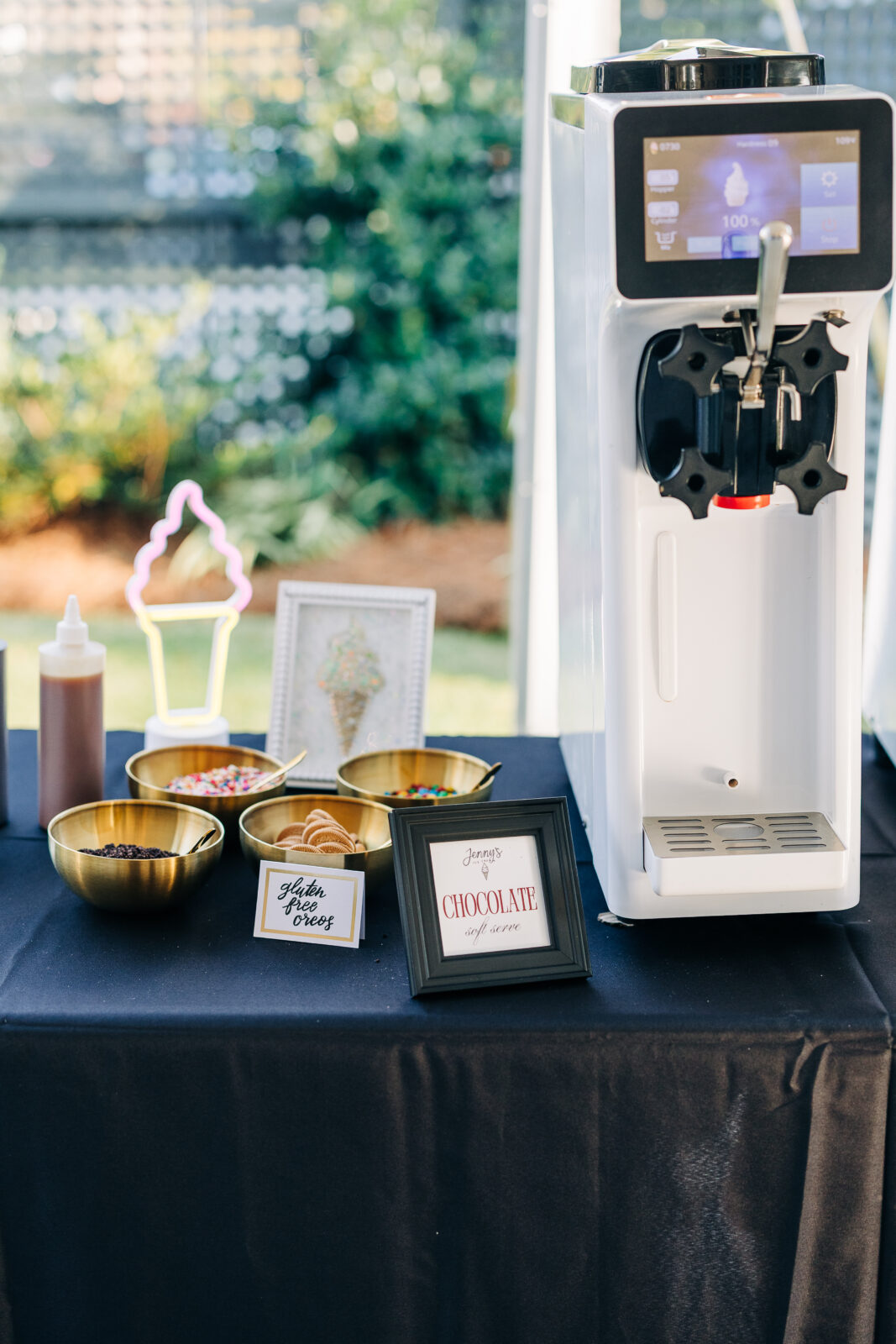 Close-up of the DIY soft-serve ice cream bar setup with a machine, bowls of toppings, and signs for "chocolate soft serve" and "gluten-free cones" at the Hartley House Wedding South Carolina reception