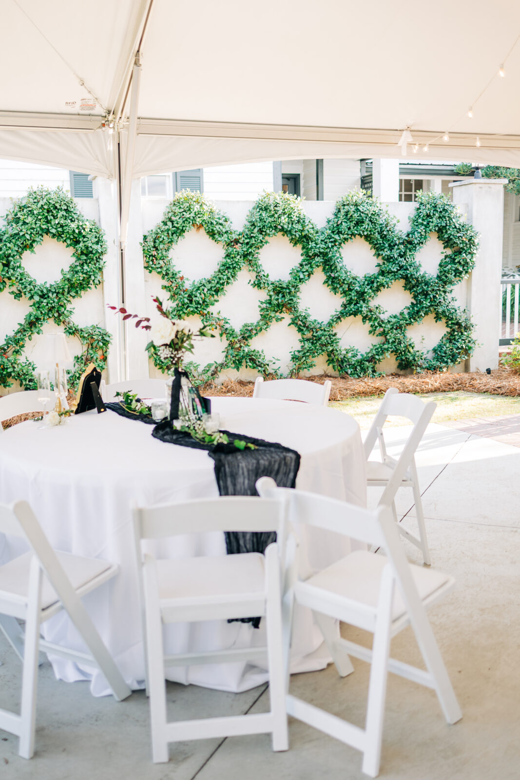 Outdoor reception setup under a white tent showing a round table with a white cloth, a black gauze runner, white chairs, and the diamond vine wall in the background at the Hartley House Wedding South Carolina