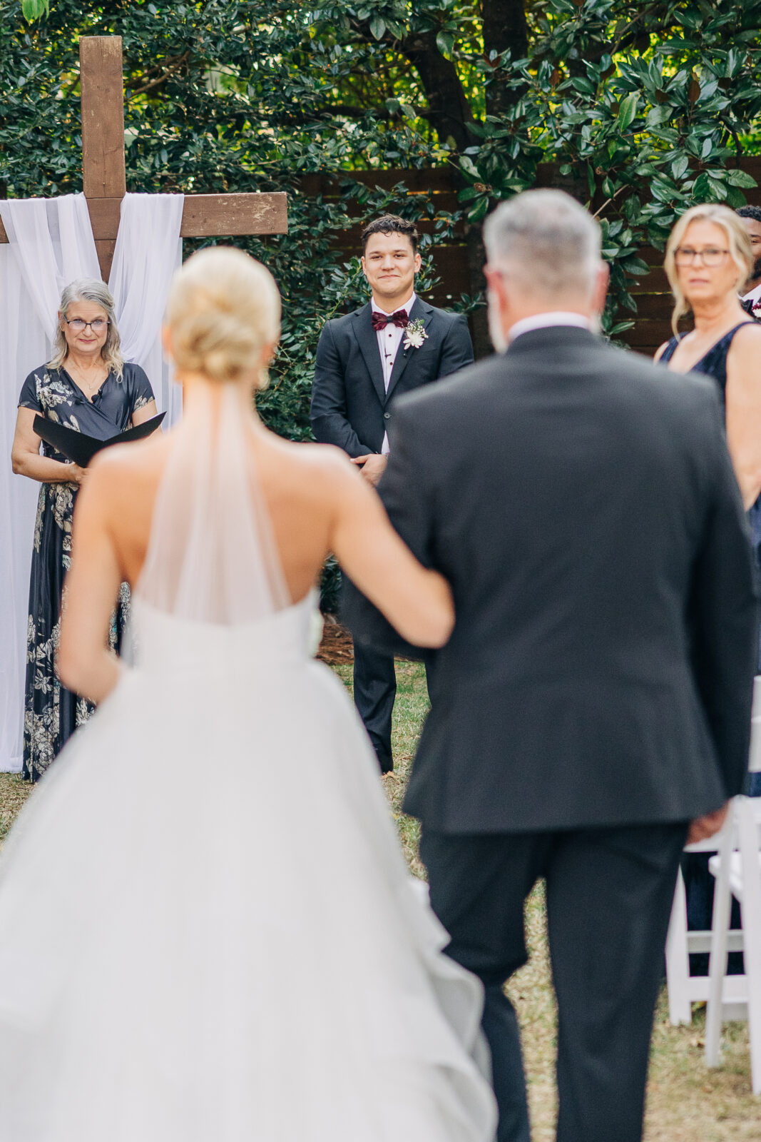 The bride, in a backless dress and veil, walks down the aisle arm-in-arm with her father toward the groom during the outdoor ceremony at the Hartley House Wedding South Carolina
